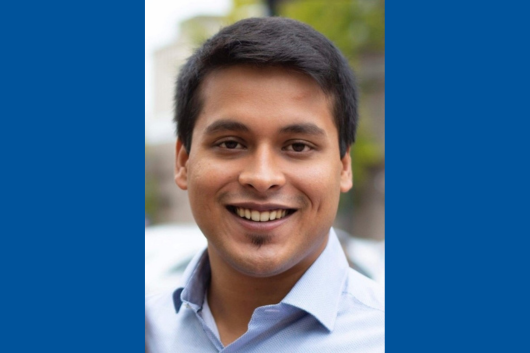 A photo of Rohit Roy, a young man with short black hair, smiling warmly at the camera. He is wearing a light blue collared shirt. The background is slightly blurred, showing an outdoor setting with greenery and buildings in the distance.